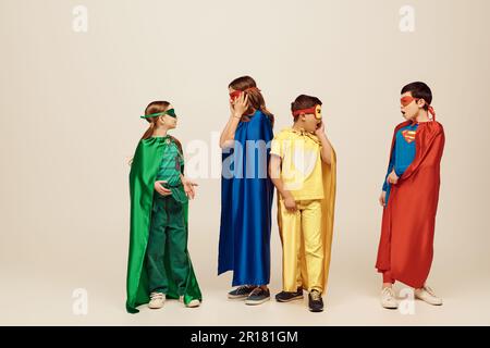 Bambini interrazziali scioccati in costumi colorati supereroi con mantello e maschere che si guardano l'un l'altro su sfondo grigio in studio, CH Internazionale Foto Stock