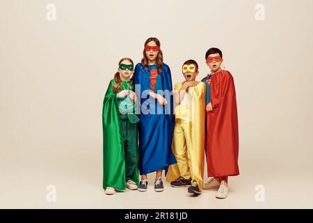 Scioccato multiculturale e preteen bambini in colorati costumi supereroi con mantello e maschere guardando la macchina fotografica su sfondo grigio in studio, Internatio Foto Stock