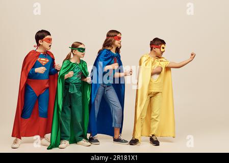 Coraggiosi bambini multiculturali in costumi colorati con mantello e maschere in piedi con pugni clenched insieme su sfondo grigio in studio, Child Pro Foto Stock