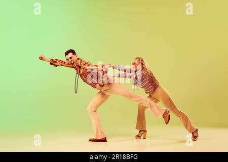 Giovane coppia espressiva e talentuosa, uomo e donna in eleganti costumi vintage che ballano su sfondo giallo verde sfumato Foto Stock