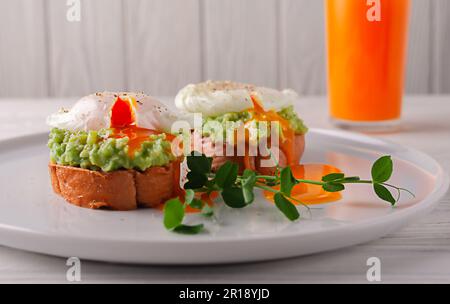 Pane tostato con avocado, uova in camicia e succo di frutta su un tavolo di legno bianco. alimentazione sana. Colazione europea. Foto Stock