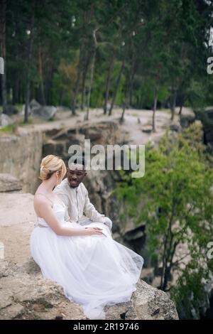 Giovani sposi novelli di coppia interrazziali siedono sulla roccia e parlano sullo sfondo della foresta e del canyon. Concetto di relazioni d'amore e di unità tra dif Foto Stock