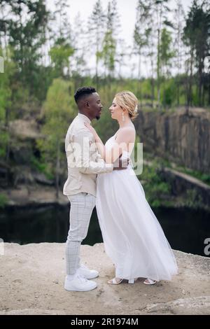 Sposi novelli felici e interrazziali si ergono sulla roccia e abbracciano sullo sfondo del lago, della foresta e del canyon. Concetto di relazioni d'amore e di unità Foto Stock