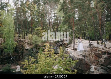 Sposi novelli felici e interrazziali si ergono sulla roccia e abbracciano sullo sfondo della foresta e del canyon. Il concetto di relazioni d'amore e di unità betwee Foto Stock