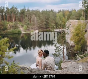 Sposi sposi novelli felici interrazziali siedono sulla roccia e abbracciano sullo sfondo bello del lago, della foresta e del canyon. Concetto di relazioni d'amore An Foto Stock