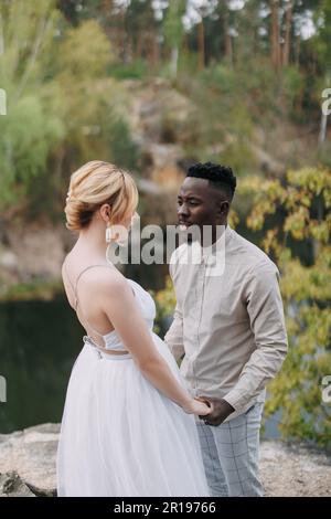 Sposi novelli di coppia interrazziali felici si leva in piedi sulla roccia e tiene le mani l'un l'altra contro lo sfondo del lago e della foresta. Concetto di relazioni d'amore e. Foto Stock