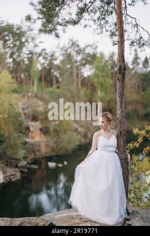 Giovane sposa felice in vestito bianco si erge sulla roccia vicino al pino sullo sfondo del lago, della foresta e del canyon. Foto Stock