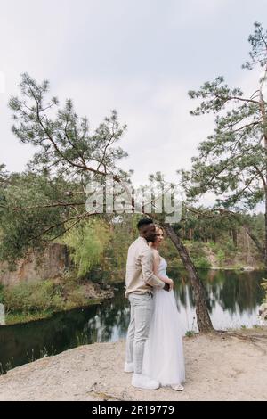 Sposi novelli felici e interrazziali si ergono sulla roccia e abbracciano sullo sfondo del lago, della foresta e del canyon. Concetto di relazioni d'amore e di unità Foto Stock