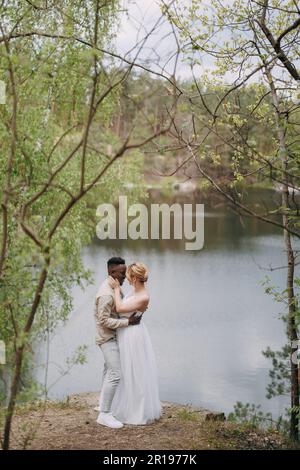 Sposi novelli di coppia interrazziali felici si leva e abbraccia sullo sfondo del lago e della foresta. Concetto di relazioni d'amore e di unità tra le differenze Foto Stock