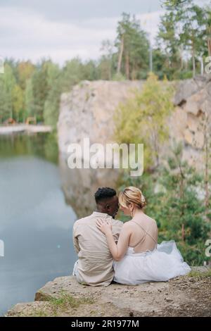 Sposi sposi novelli felici interrazziali siedono sulla roccia e abbracciano sullo sfondo bello del lago, della foresta e del canyon. Concetto di relazioni d'amore An Foto Stock