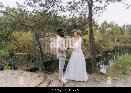 Sposi novelli di coppia interrazziali felici si leva e tiene le mani contro lo sfondo del lago e della foresta. Concetto di relazioni d'amore e di unità essere Foto Stock