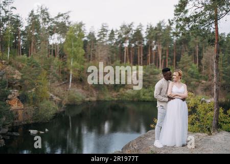 Sposi novelli felici e interrazziali si ergono sulla roccia e abbracciano sullo sfondo del lago, della foresta e del canyon. Concetto di relazioni d'amore e di unità Foto Stock