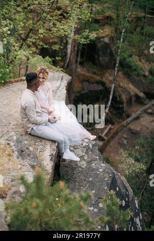 Giovani sposi novelli di coppia interrazziali siedono sulla roccia e parlano sullo sfondo della foresta e del canyon. Concetto di relazioni d'amore e di unità tra dif Foto Stock