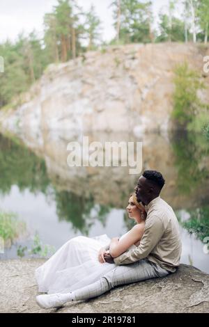 Sposi sposi novelli felici interrazziali siedono sulla roccia e abbracciano sullo sfondo bello del lago, della foresta e del canyon. Concetto di relazioni d'amore An Foto Stock