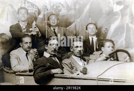 Divertente cartolina fotografica vintage di 7 uomini di fronte a uno sfondo guida una 'auto' - alcuni stanno mangiando coni gelato. Foto Stock