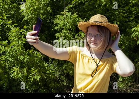 Ragazza asiatica in occhiali, cappello di paglia e vestito giallo spara video sul suo telefono, prende selfie, comunica online, blogger Foto Stock