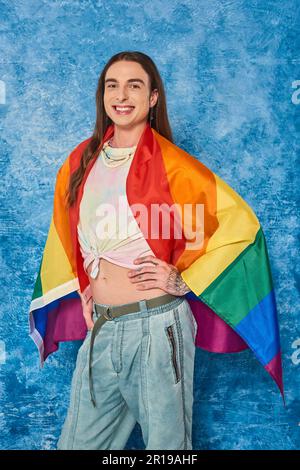 Felice uomo gay con capelli lunghi e tatuati in abiti casual con bandiera lgbt posa e guardare la macchina fotografica mentre festeggia il mese di orgoglio su bac blu chiazzato Foto Stock