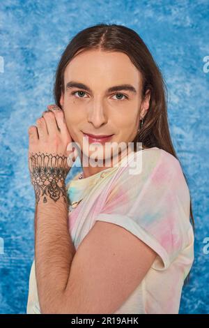 Ritratto di giovane uomo gay dai capelli lunghi e tatuati che guarda la macchina fotografica durante la comunità lgbt e la celebrazione del mese di orgoglio su sfondo blu chiazzato Foto Stock