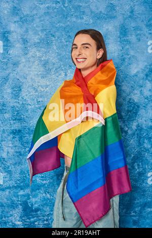 Uomo omosessuale positivo a capelli lunghi che guarda e fotocamera e che si avvolge nella bandiera lgbt durante la celebrazione del mese della comunità di orgoglio su sfondo blu chiazzato Foto Stock