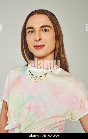 Ritratto di giovane e uomo omosessuale a capelli lunghi in tie dye t-shirt guardando la macchina fotografica isolato sul grigio, felice orgoglio comunità mese celebrazione concep Foto Stock