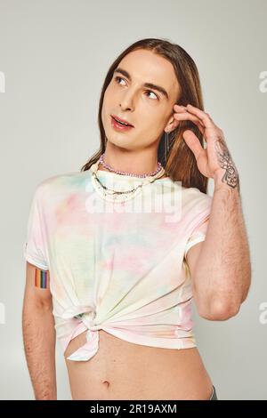 Uomo omosessuale dai capelli lunghi e sognanti in T-shirt tie dye che tocca i capelli e guarda via durante la celebrazione del mese di orgoglio lgbt mentre si trova isolato sopra Foto Stock