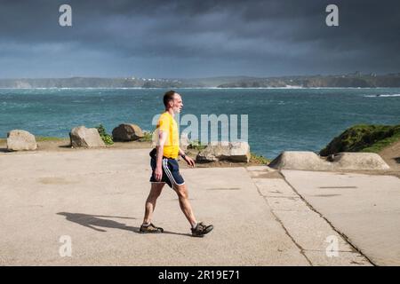 Un'immagine panoramica di un uomo che indossa pantaloncini a piedi lungo la strada costiera su Towan Head che si affaccia sulla Newquay Bay in Cornovaglia nel Regno Unito. Foto Stock