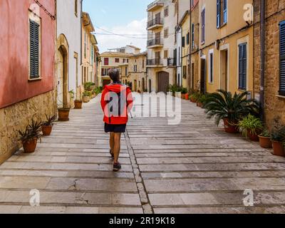 Donna in giacca arancione che cammina per le graziose strade di Alcudia, una città fortificata sulla costa nord-orientale di Maiorca Spagna Foto Stock