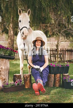 Donna in abiti da lavoro, guanti, stivali di gomma e cappello di paglia seduto vicino a vasi di fiori in giardino, bianco cavallo arabo vicino, alberi verdi sfocati e cortile b Foto Stock
