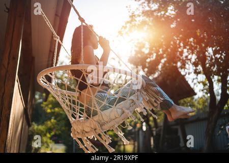 felice giovane donna cavalcata su sedia altalena macrame vicino a casa di campagna all'aperto Foto Stock