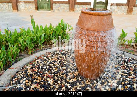 Una bella fontana a forma di vaso marrone, una brocca con gocce d'acqua cadenti su pietre colorate in piedi in un letto di fiori con pianta verde Foto Stock