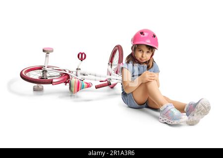 Bambina che indossa una bicicletta che tiene il ginocchio ferito e che si siede a terra isolata su sfondo bianco Foto Stock