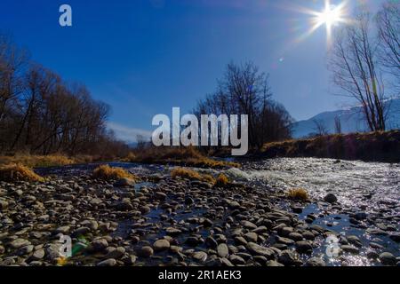 Pietre di ghiaia che rotolano su un ampio paesaggio fluviale in una foresta senza fronde d'inverno sotto un cielo azzurro soleggiato con sole che splende Foto Stock