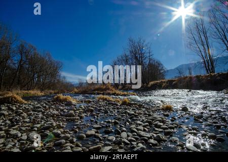 Pietre di ghiaia che rotolano su un ampio paesaggio fluviale in una foresta senza fronde d'inverno sotto un cielo azzurro soleggiato con sole che splende Foto Stock