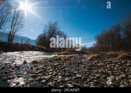 Pietre di ghiaia che rotolano su un ampio paesaggio fluviale in una foresta senza fronde d'inverno sotto un cielo azzurro soleggiato con sole che splende Foto Stock