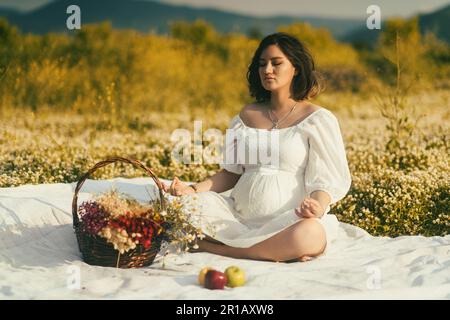 Seduto a gambe incrociate su una coperta da picnic bianca con gli occhi chiusi, una donna incinta medita tra margherite, un cesto di fiori secchi, e mele vicino Foto Stock