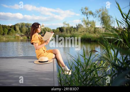 Giovane donna che legge libro vicino al lago nelle giornate di sole Foto Stock