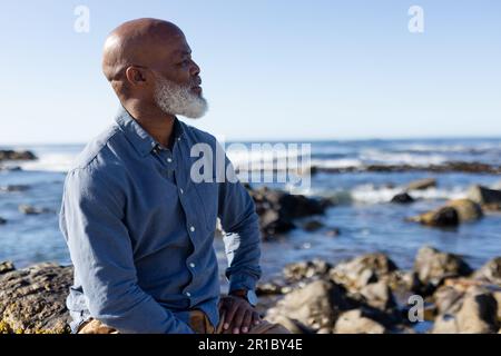 Uomo afroamericano attento seduto sulle rocce e ammirando la vista sul mare Foto Stock