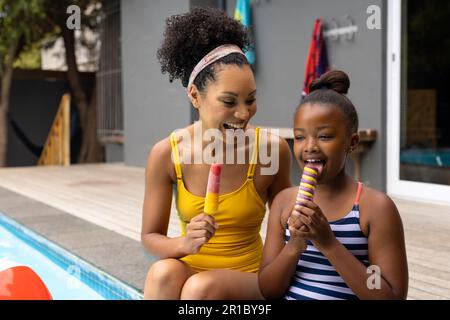 Buona madre afro-americana e figlia seduta in piscina in costume da bagno che mangia gelati Foto Stock