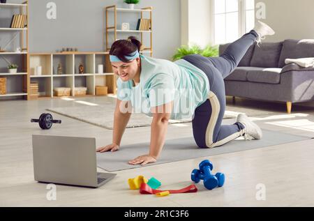 Donna felice in sovrappeso che usa il laptop e fa l'allenamento sportivo con un allenatore di fitness remoto Foto Stock