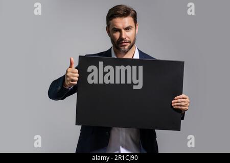 Uomo serio con i pollici in su e la targhetta vuota. Copiare spazio per il testo o il disegno. Bell'uomo che mostra una scheda pubblicitaria vuota sullo sfondo dello studio. Foto Stock