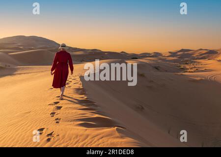 Giovane bella donna in vestito rosso cammina lungo dune di sabbia raggiungendo la cima da sola Foto Stock