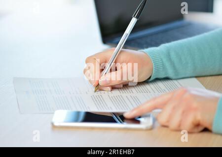 Primo piano di una donna che scrive a mano o firma un documento sulla consultazione di un telefono cellulare su una scrivania a casa o in ufficio Foto Stock