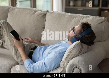 Uomo giovane e rilassato che indossa cuffie wireless e si appoggia sul divano di casa Foto Stock
