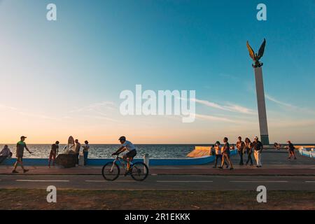 I residenti si rilassano lungo la malecón di fronte alla scultura dell'Angelo Maya a Campeche, Messico, al tramonto Foto Stock