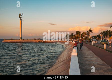 I residenti si rilassano lungo la malecón di fronte alla scultura dell'Angelo Maya a Campeche, Messico, al tramonto Foto Stock