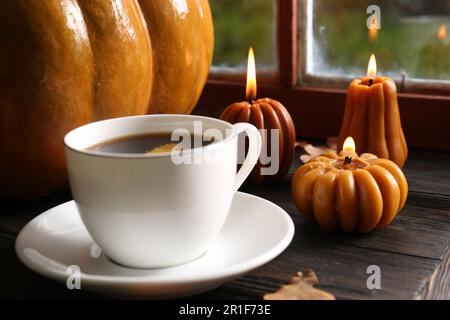 Tazza di bevanda calda e candele a forma di zucca sul tavolo di legno vicino alla finestra, primo piano. Accogliente atmosfera autunnale Foto Stock