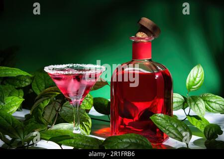 Cocktail rosso con ghiaccio in un bicchiere di margarita con bordo di zucchero, accanto ad esso si trova l'alcol rosso in bottiglia con tappo di legno. Circondato da foglie verdi di Foto Stock