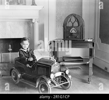 Foto di archivio: Giovane ragazzo che gioca in un'automobile giocattolo in un salotto, con radio ca. 1910s o 1920s Foto Stock