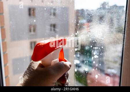 spruzzare liquido di lavaggio sulla superficie di vetro vicino durante la pulizia della finestra di casa in alta casa di città il giorno di primavera Foto Stock