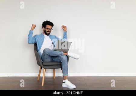 Gioioso indiano maschio utilizzando il computer portatile e scuotendo Fists mentre si siede in sedia Foto Stock